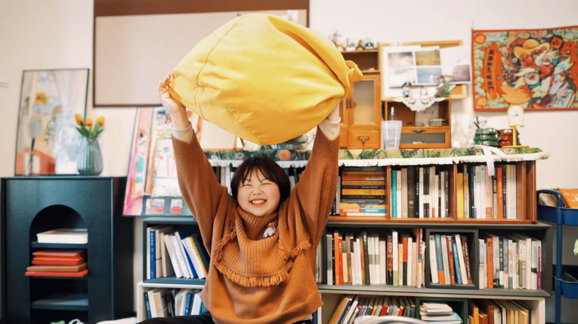 Jundao Hou smiling and lifting a bright yellow cushion in a creative studio